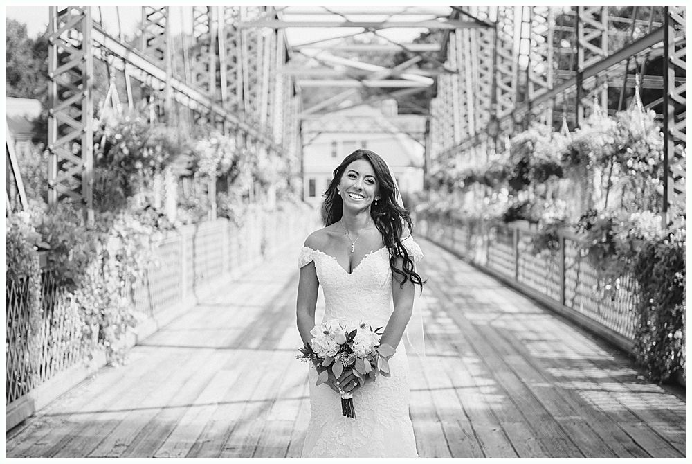 Bride in a white gown holding flowers, standing on a bridge, smiling.