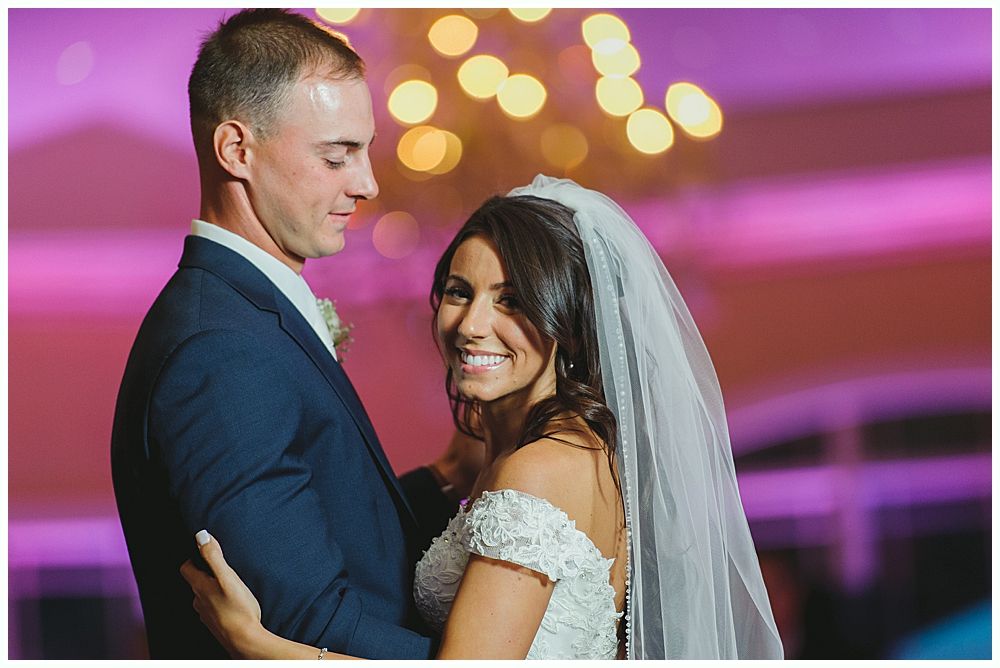 Bride and groom dancing, bride smiles, groom looks at her. Indoors with soft lighting.