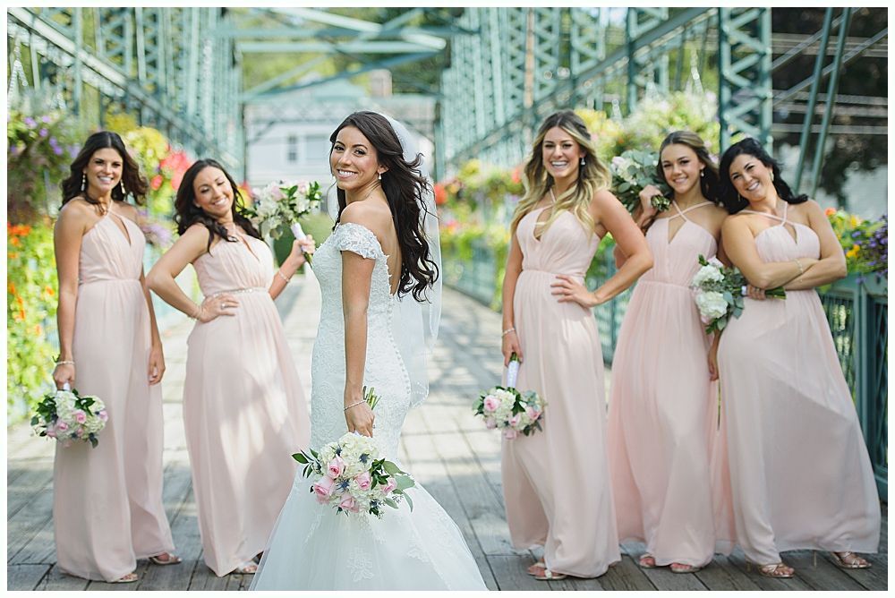 Bride with bridesmaids on a bridge, all wearing pink dresses, holding bouquets, and smiling.