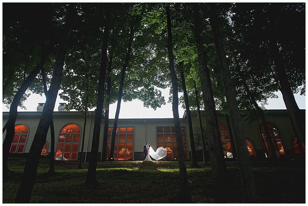 Couple in wedding attire stands before a building with orange windows, framed by trees.