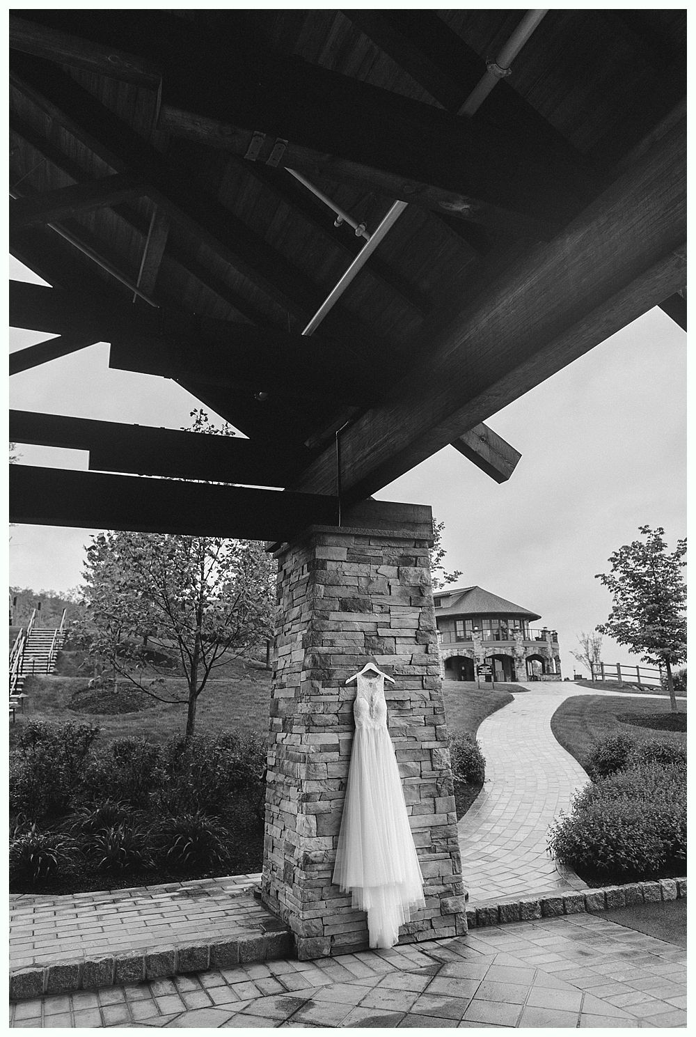 Wedding dress hanging on a stone pillar under a wooden structure, with a paved path leading to a building.