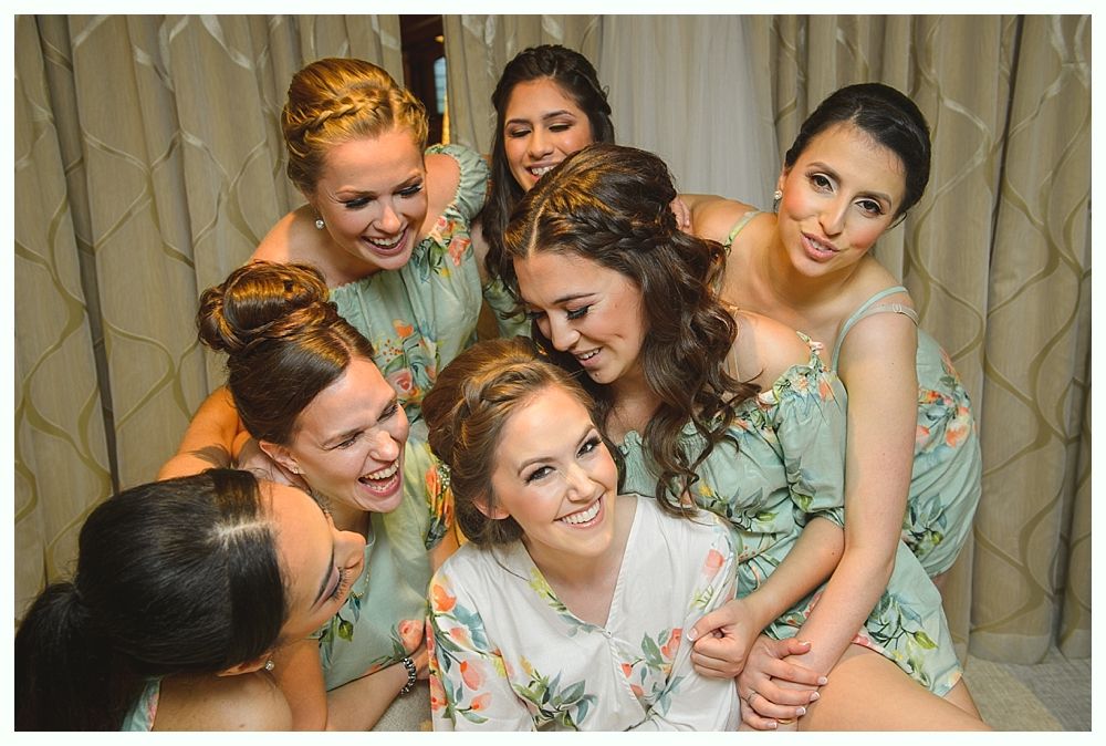 Bridesmaids surround a bride, laughing. They wear floral robes, posing in a room with neutral curtains.