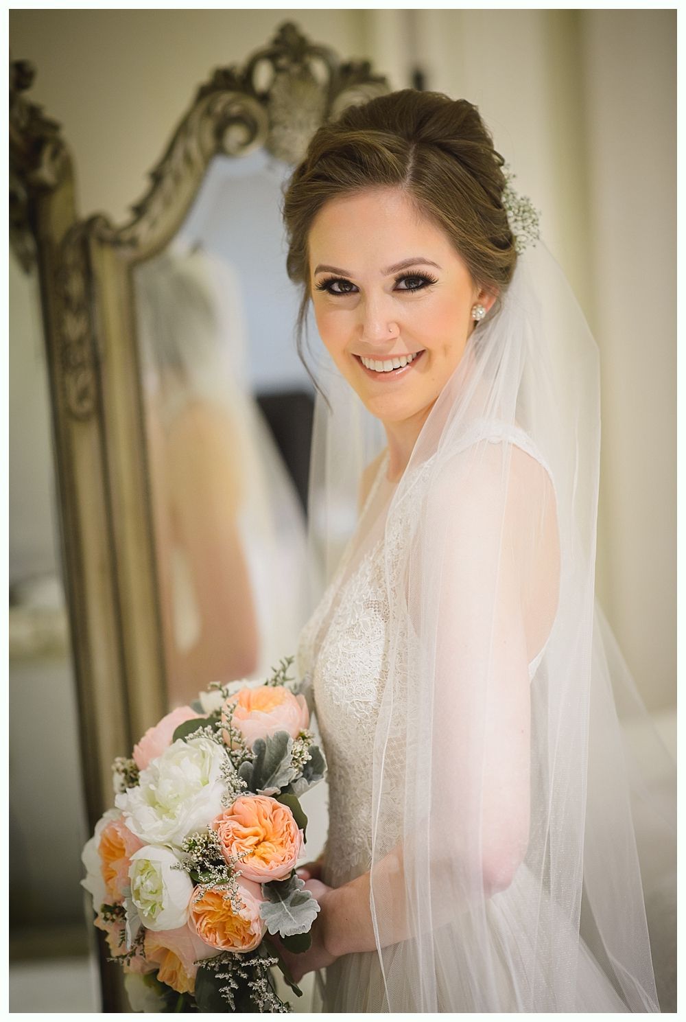Bride holding bouquet, smiling near a mirror. She wears a veil and a beaded gown.