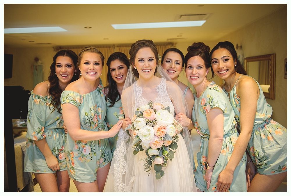 Bride in white dress with bouquet, surrounded by bridesmaids in floral-print rompers, smiling in a room.
