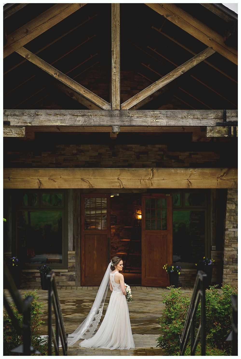 Bride in wedding dress, holding bouquet, standing on steps in front of wooden building with open doors.