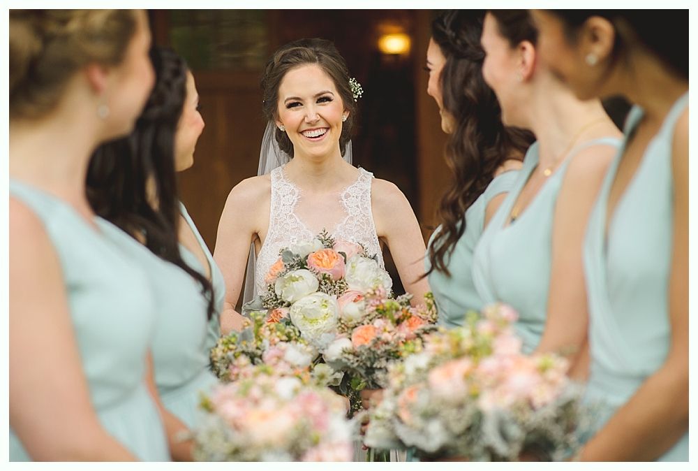 Bride in white lace dress smiles, surrounded by bridesmaids in light blue dresses, holding bouquets.