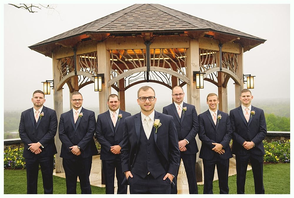 Groom and groomsmen in navy suits stand in front of a wooden gazebo on a cloudy day.