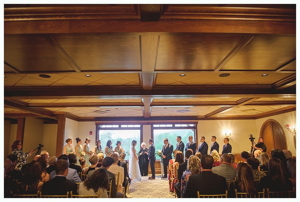 Wedding ceremony indoors, couple at altar, guests seated, windows behind.