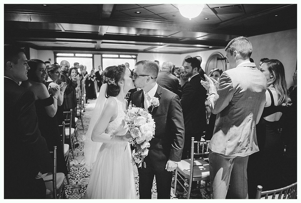 Bride and groom kiss, cheered by wedding guests. Ceremony in a room with windows and chandeliers.