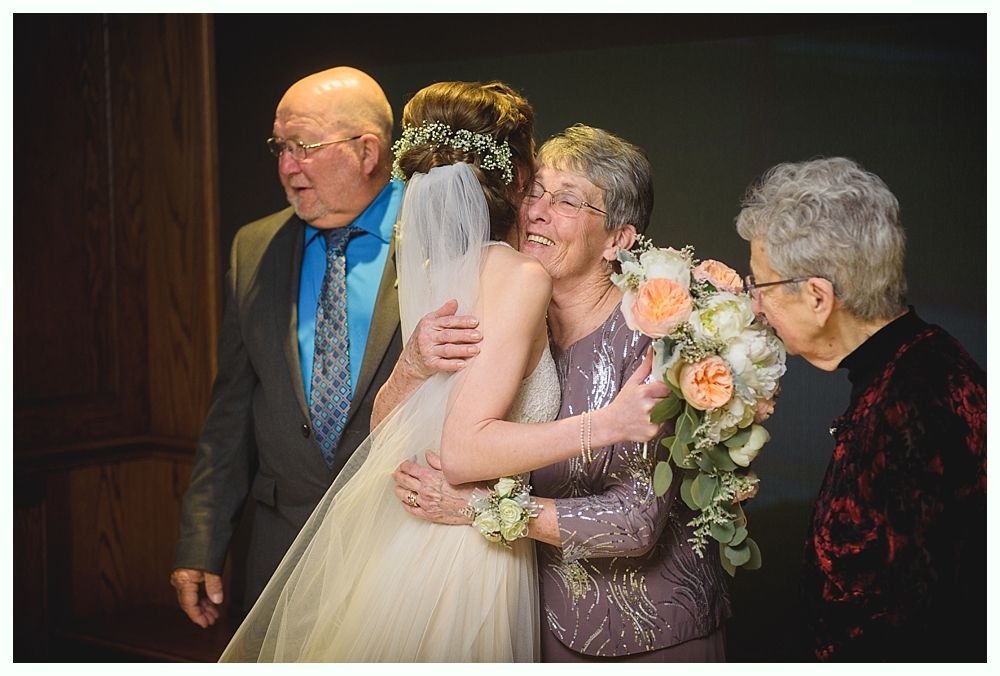 Bride hugs two women, while a man looks on. One woman sniffs the bride's bouquet. Indoor setting, soft lighting.