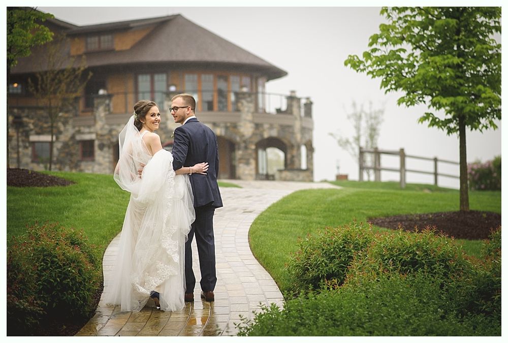 Bride and groom walk hand in hand down a paved path in front of a stone building with green landscaping.