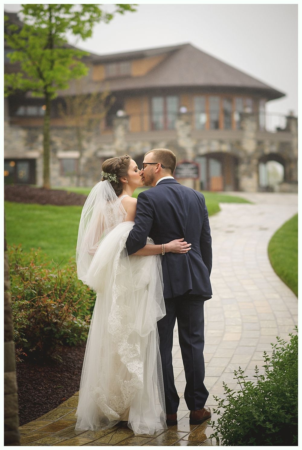Bride and groom kissing outdoors in front of stone building. Bride in white dress, groom in blue suit.