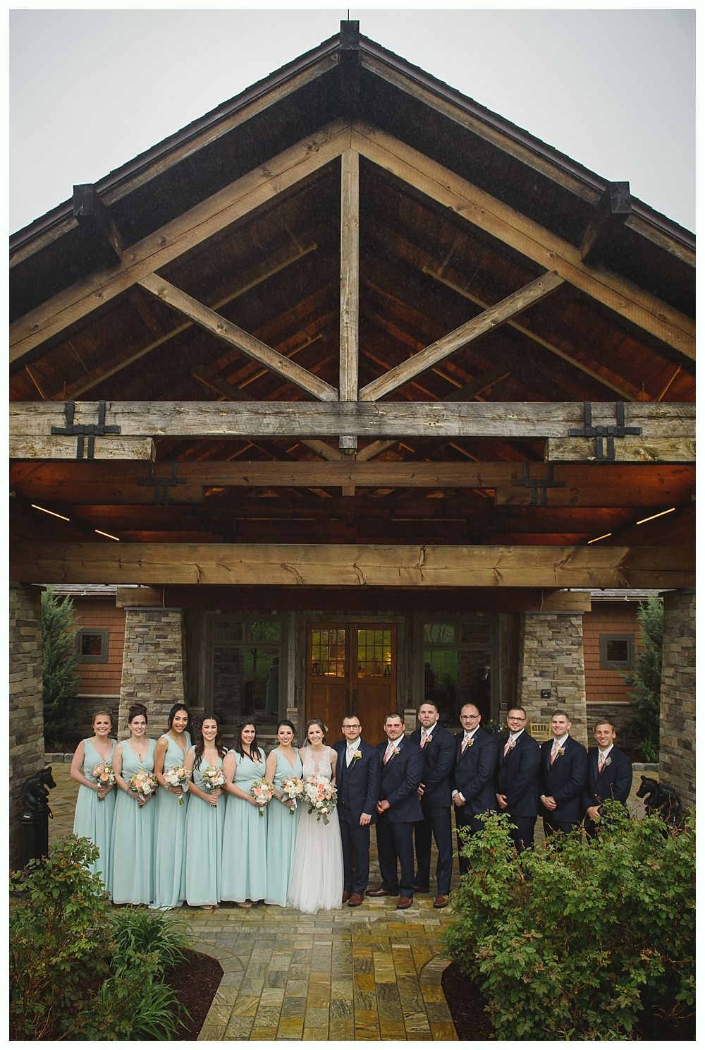 Wedding party in teal dresses and navy suits posing outside a wooden building with a stone facade.