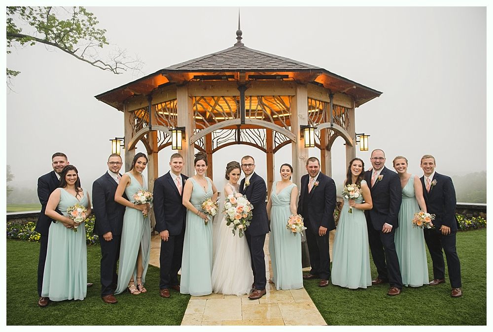 Wedding party poses in front of a gazebo. Bridesmaids wear teal dresses. Groomsmen wear navy suits. Cloudy day.