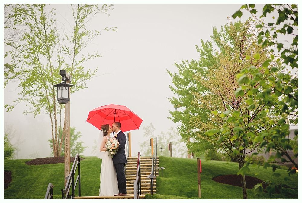 Couple kissing under a red umbrella on a rainy day, outdoors by steps and trees.