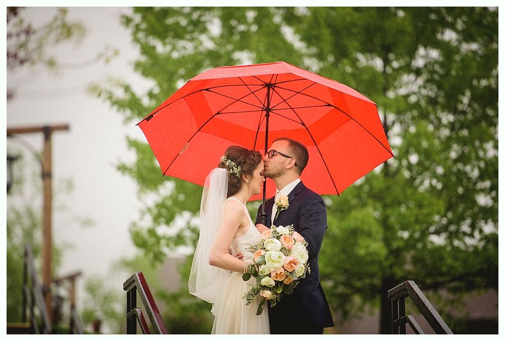 Bride and groom under a red umbrella, kissing. The setting is outdoors, with greenery in the background.
