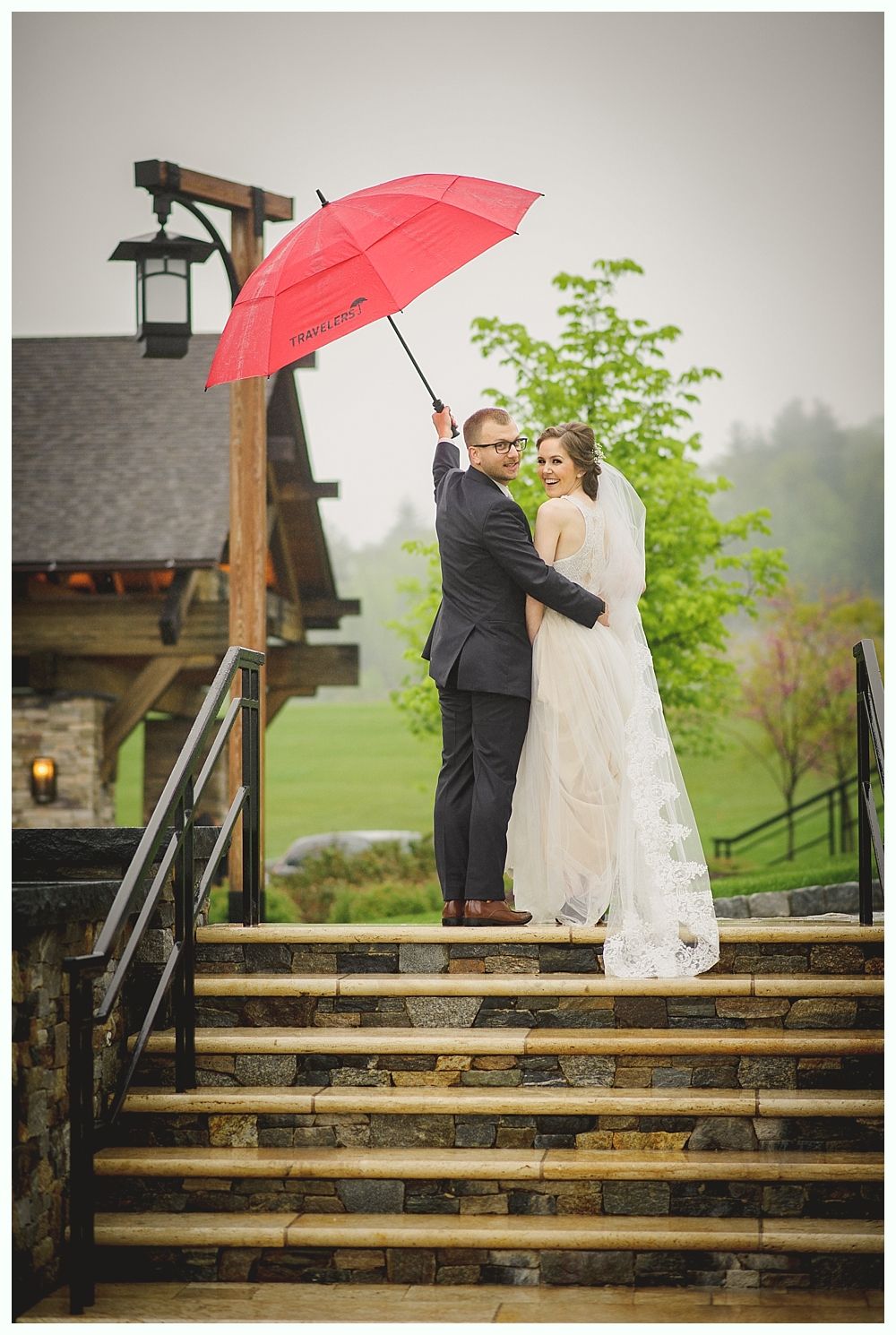 Newlyweds embrace on stone steps under a red umbrella, in the rain.