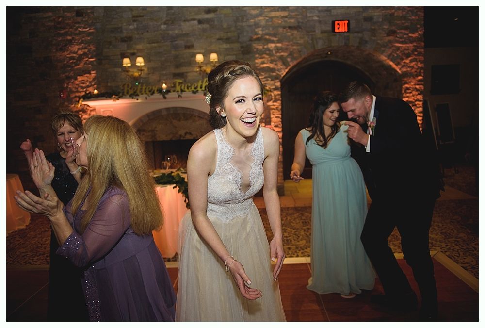 Bride laughing, dancing at a reception, with guests clapping and a groom laughing nearby. Warm-toned interior.