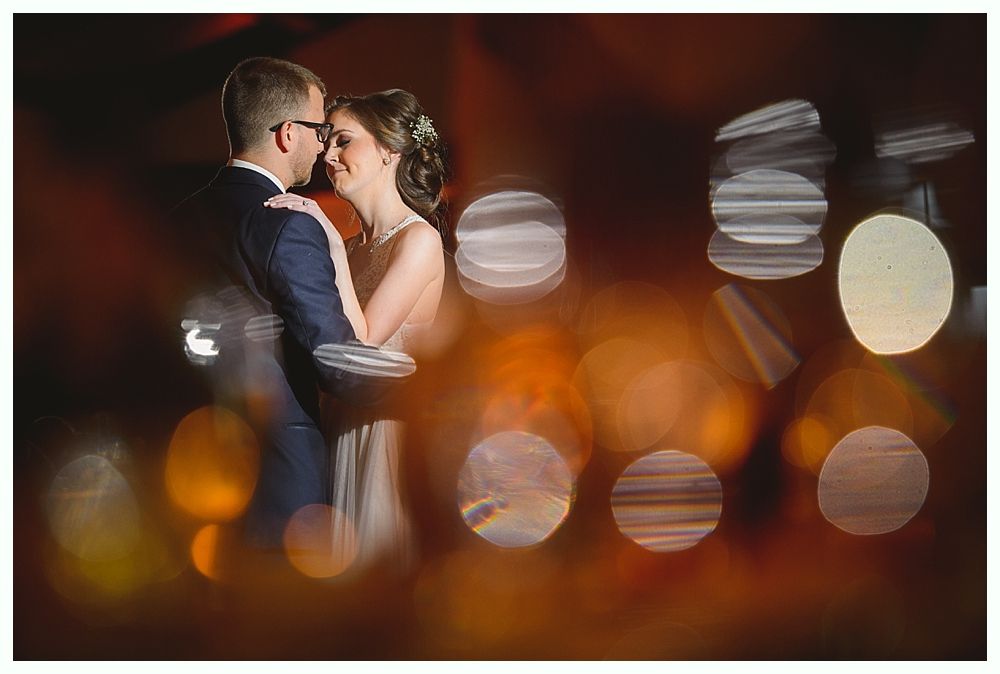 Couple embracing, blurred lights in foreground, wedding attire. Warm tones.