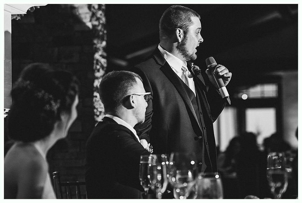 Man in suit giving a speech with a microphone at a wedding reception.