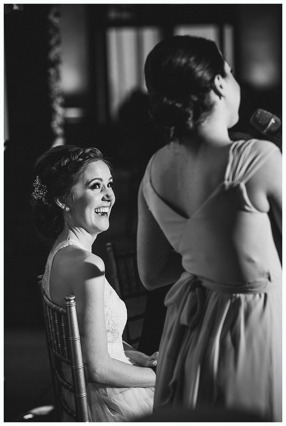 Bride smiles while a bridesmaid speaks into a microphone at a wedding reception.