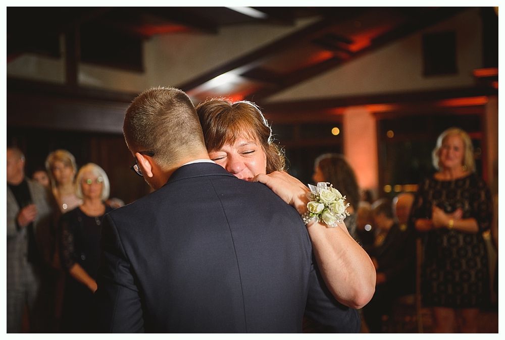 A man in a suit dances and hugs a woman; she has a bouquet and looks emotional. Indoor setting, soft lighting.