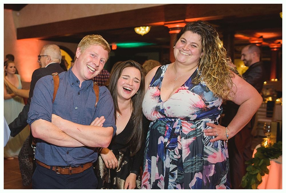 Three people laughing at a party: a man with arms crossed, and two women in floral print. Dim interior.