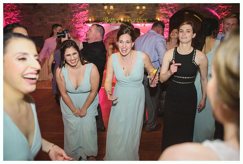 People dancing at an event, wearing gowns. Some are smiling and laughing, on a wooden floor, with pink lighting.