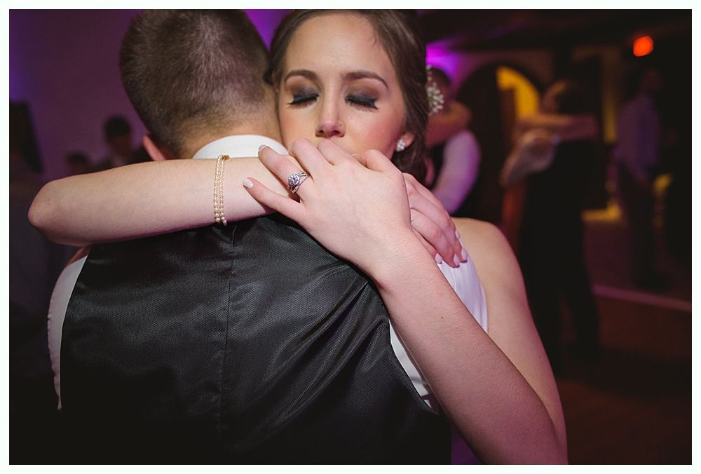 Bride hugs groom tightly, eyes closed, on dance floor during reception.