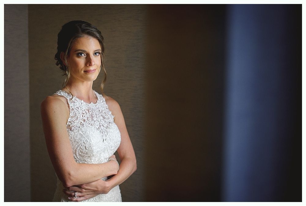 Bride in a white lace dress, arms crossed, looking at the camera, standing by a window with natural light.