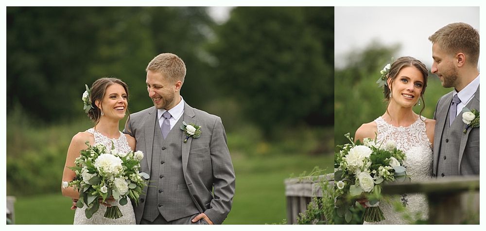 Newlyweds smiling, embracing outdoors; green backdrop. Woman in white lace dress, man in gray suit.