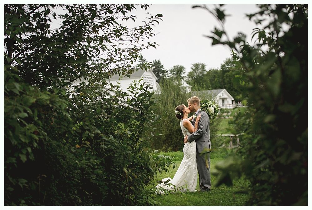 Couple kisses in garden with white buildings in background.