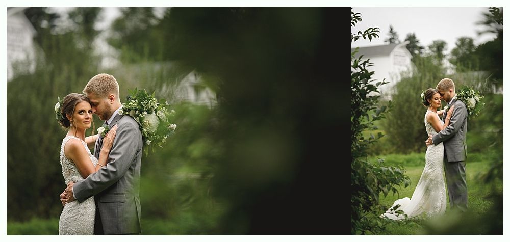 A newlywed couple embraces outdoors. The bride wears a white dress. The groom wears a gray suit. They are surrounded by greenery.