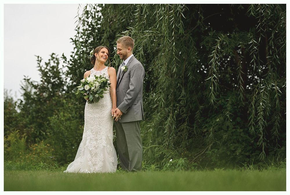 Bride and groom holding hands, smiling. She wears a white lace dress and holds flowers. He wears a gray suit, standing outside by a tree.
