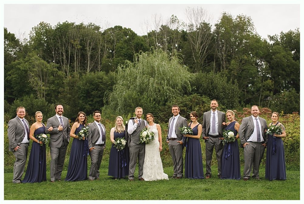Wedding party poses in a field, bride in white, bridesmaids in navy, groomsmen in gray suits, green trees in background.