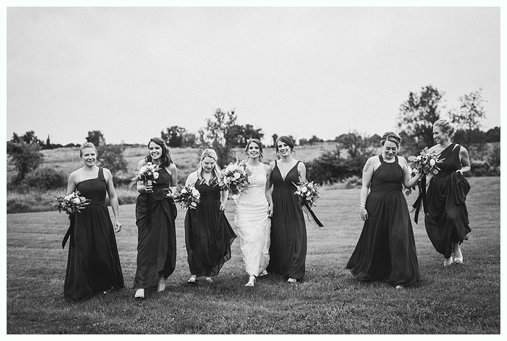 Bride and bridesmaids walking through a field, smiling, holding bouquets. Black and white photo.
