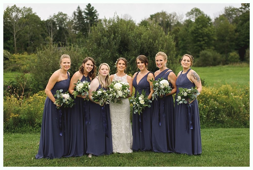 Bride poses with bridesmaids in navy gowns, holding white bouquets, outdoors.