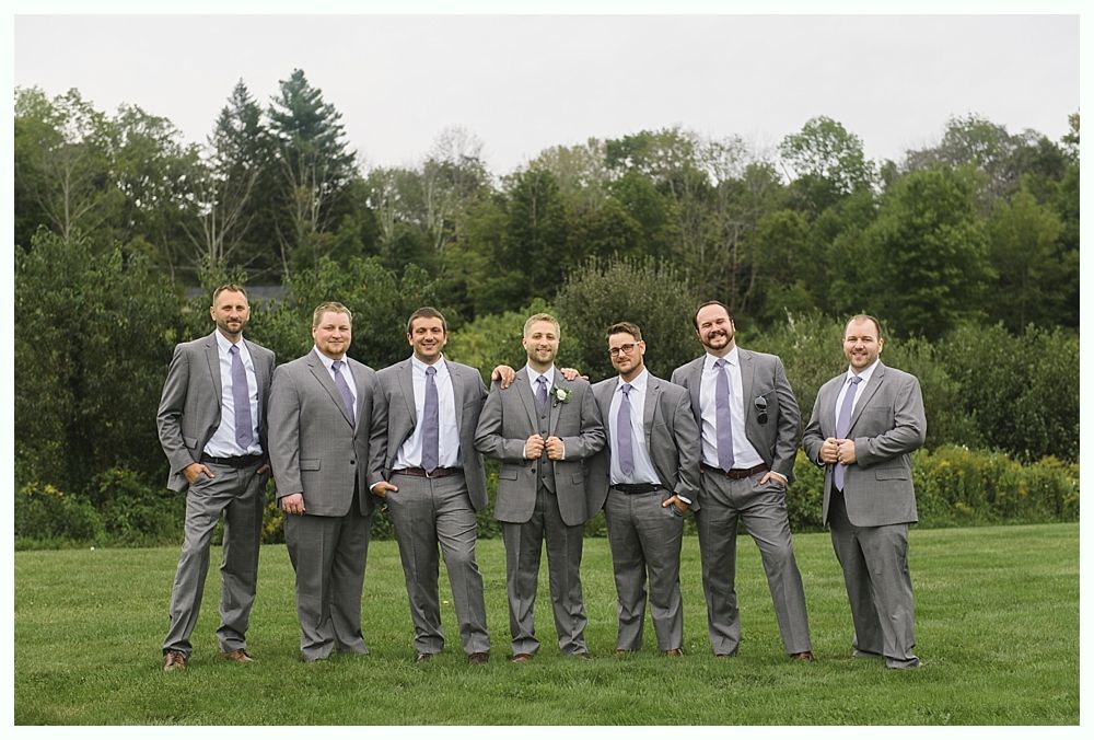 Seven men in gray suits and purple ties pose in a grassy field with trees in the background.