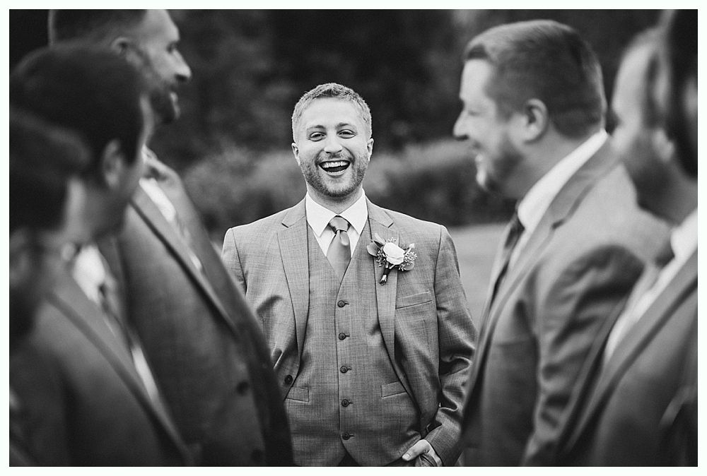 Groom laughs amidst groomsmen, all in suits, outdoors. Black and white photo.