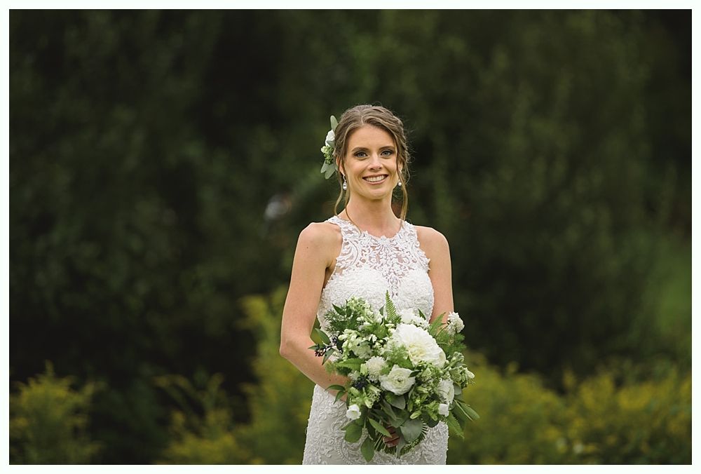 Bride in white lace dress holding bouquet, smiling outdoors against green foliage.