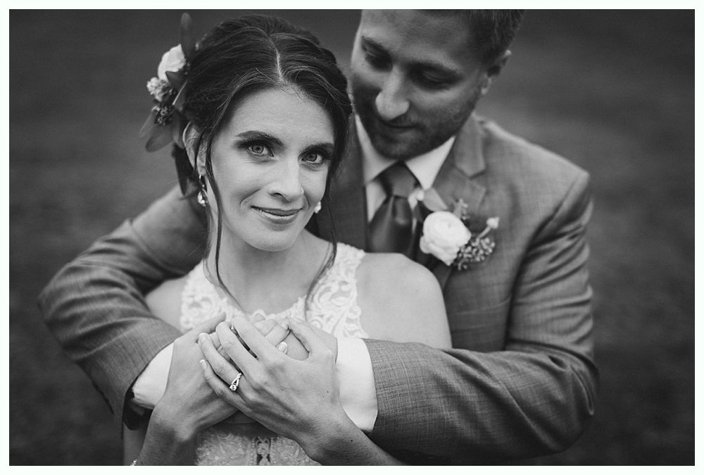 Bride and groom embrace, smiling. Black and white photo with outdoor setting.