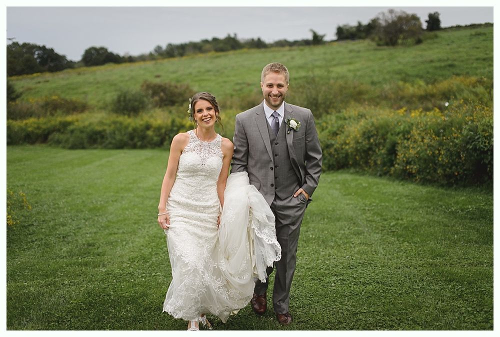 Bride and groom walk hand-in-hand in a grassy field. The bride wears a white lace dress. The groom is in a grey suit.