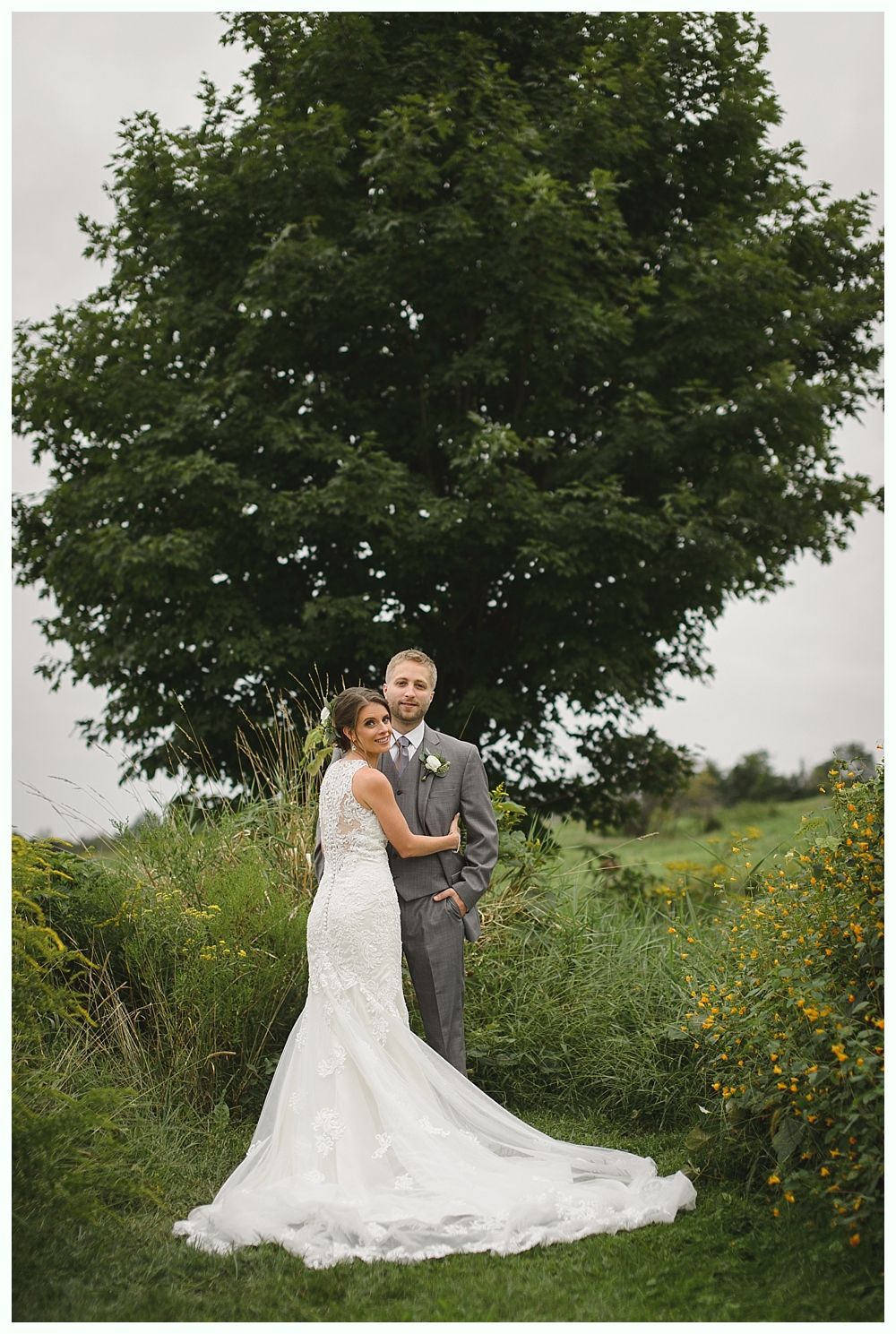 Bride and groom pose near a large tree in a field. The bride wears a white lace gown; the groom, a grey suit.