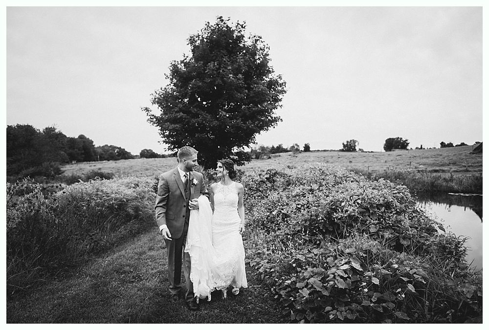 Couple in wedding attire walk in a field near a tree and a small body of water. Black and white.