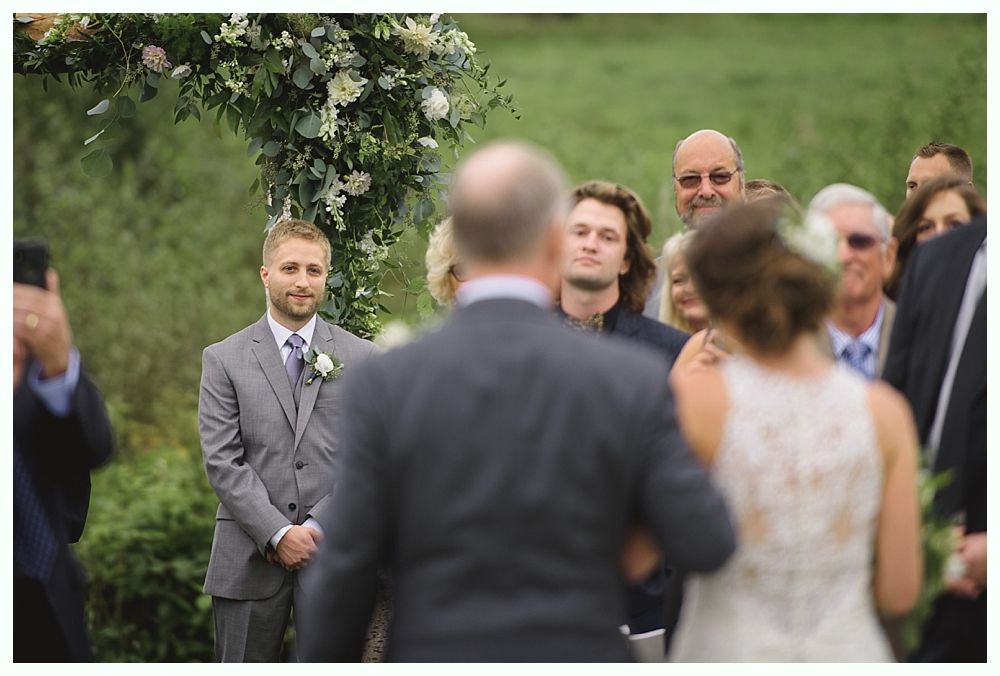 Groom waiting at the altar, looking at bride walking down the aisle, with guests and floral arch in background.