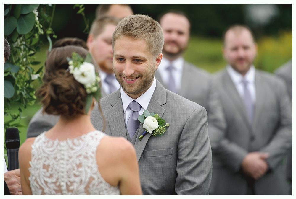 Groom smiling at bride during outdoor wedding ceremony. Men in gray suits behind.