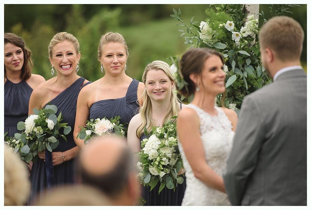 Wedding ceremony. Bride and groom face each other. Bridesmaids in navy dresses with bouquets watch the couple. Green outdoor setting.