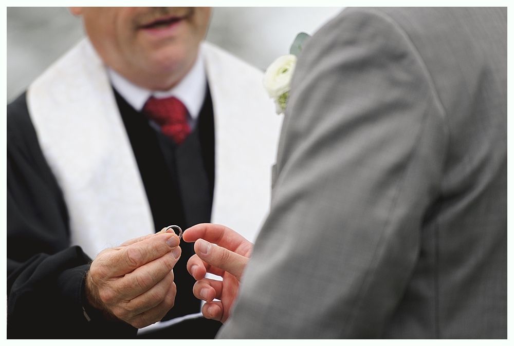 Man in robe holding wedding ring, placing it on another man's finger, outdoors.