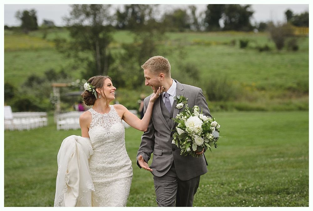 Bride and groom walking in a field. Bride touches groom's face; he smiles at her.