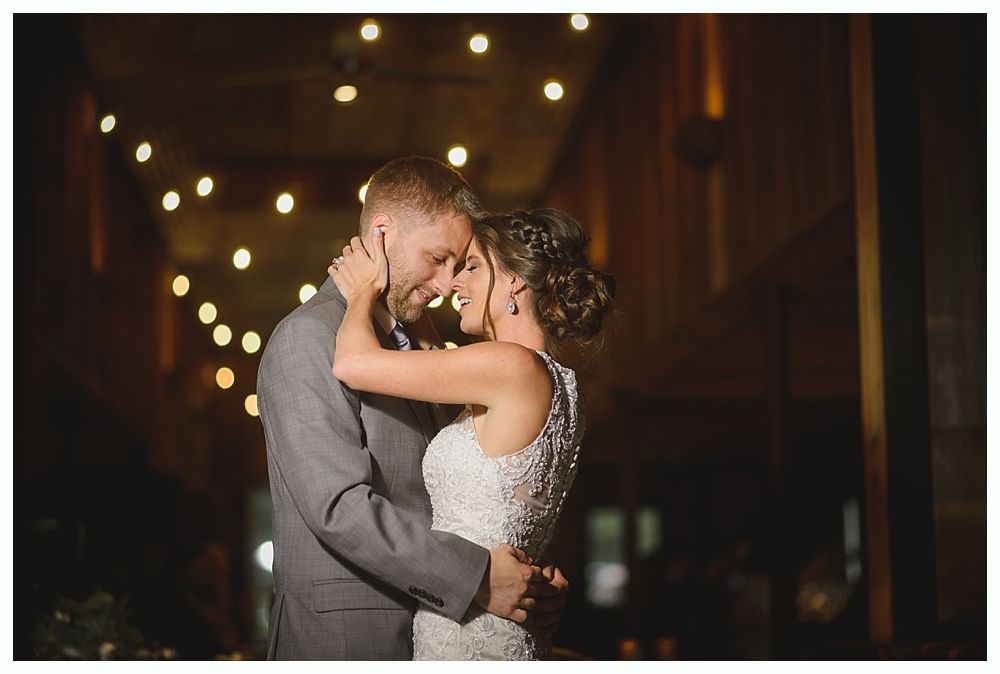 Couple embraces, lit by string lights. The bride wears a white dress, the groom a gray suit.